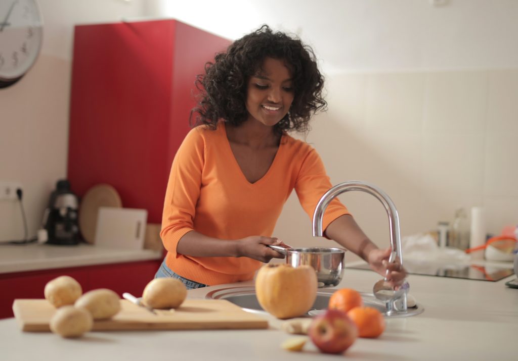 Young woman smiling as she uses kitchen sink to fill a cooking pot; food on a cutting board next to her.