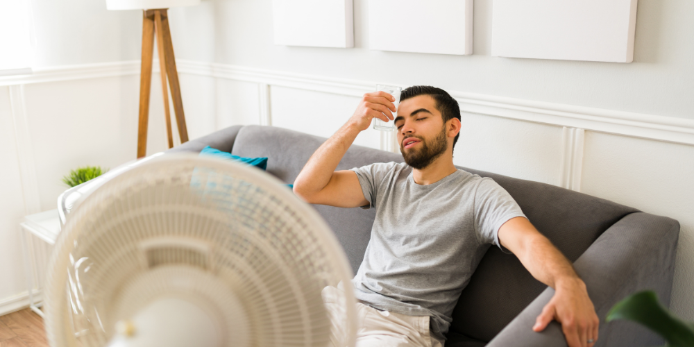 Man sitting on couch in home in front of fan, cooling his forehead with a cup of water because he's hot - Why Is My House Humid When the AC is Running - Black Diamond Plumbing & Mechanical